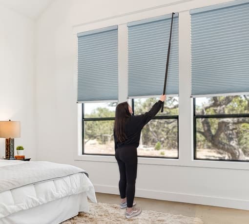 Woman adjusting blue honeycomb cellular shades with the Norman AutoWand in a bright bedroom