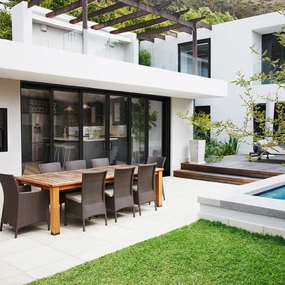 Modern white home with a pergola-shaded patio and black-framed PGT EnergyVue vinyl sliding glass doors opening to an outdoor dining area beside a pool