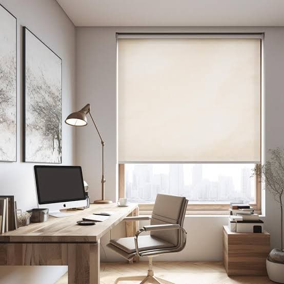 Cream Basix roller shade lowered over a large home-office window with a city view, above a wood desk and task chair