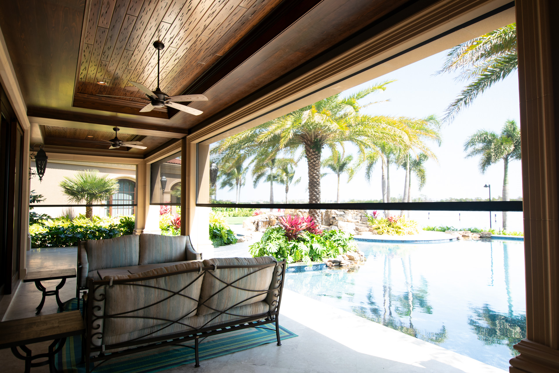 Dark Fenetex One-Track insect screens lowered around a covered lanai with a wood-slat ceiling, overlooking a resort-style pool framed by royal palms