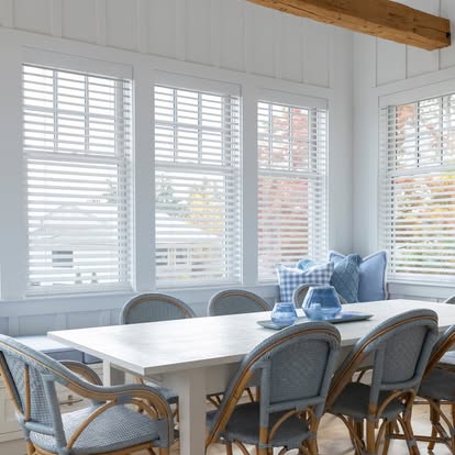 Bright coastal dining room with white plantation shutters across a bank of windows and blue woven chairs