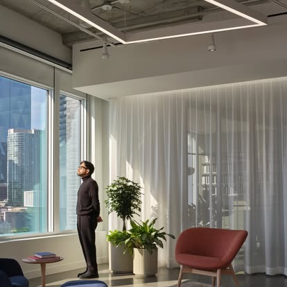Modern office corner with sheer white drapery as a person looks up at architectural lighting and a city view