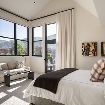 Bright bedroom with white drapery on corner windows framing a mountain-town view