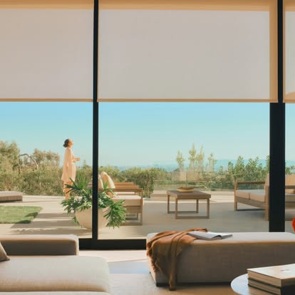 Motorized solar roller shades half-lowered over floor-to-ceiling glass as a woman stands on a poolside patio