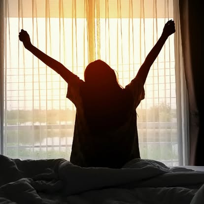 Silhouette of a woman stretching at sunrise as she opens sheer curtains over a bright window