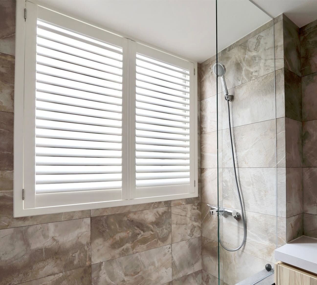 White waterproof AquaShield plantation shutters on a window inside a marble-tiled bathroom next to a glass shower