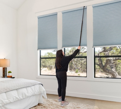 Woman adjusting blue honeycomb cellular shades with the Norman AutoWand in a bright bedroom