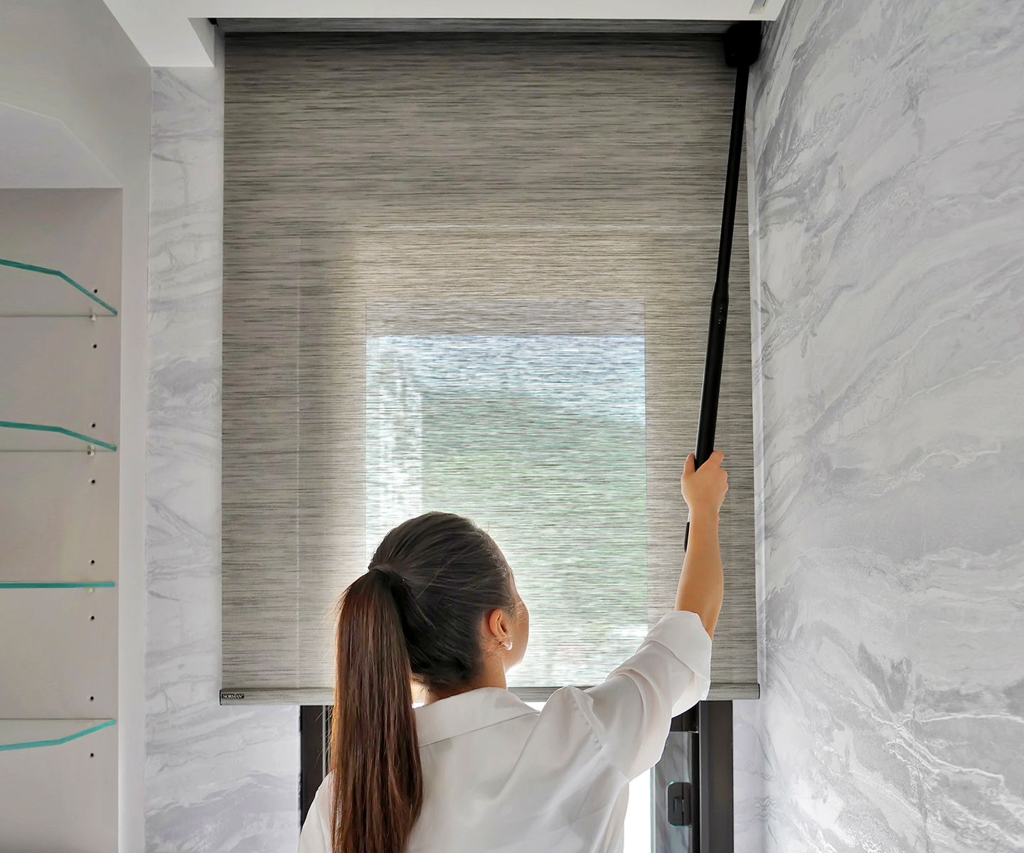 Woman raising a gray woven-texture Norman roller shade with the AutoWand charging wand in a marble-walled room