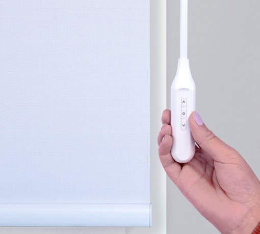 Close-up of a hand holding the Norman AutoWand charging wand with up, stop, and down buttons beside a white roller shade