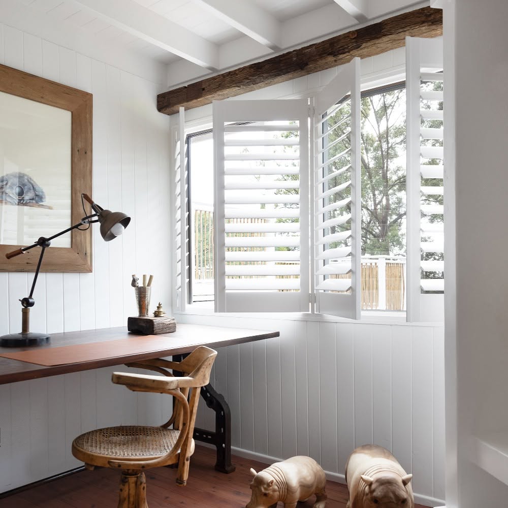 White Brightwood shutters on a corner window with one panel swung open, in a rustic-modern home office with a reclaimed wood beam and wood desk