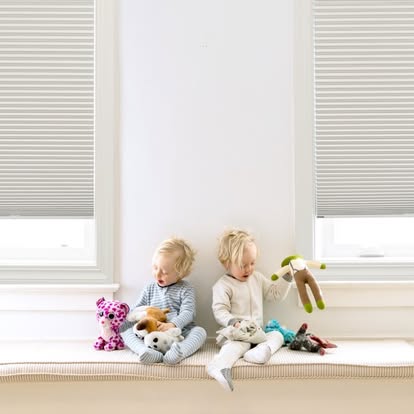 Two toddlers sitting on a window bench framed by white cordless cellular honeycomb shades