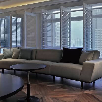 White plantation shutters wrapping floor-to-ceiling windows behind a gray sofa with a high-rise city view