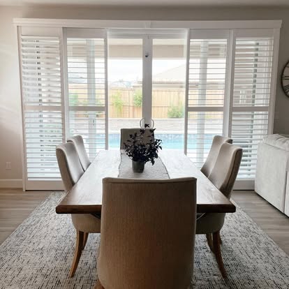Floor-to-ceiling plantation shutters across a sliding glass door in a dining room overlooking a pool