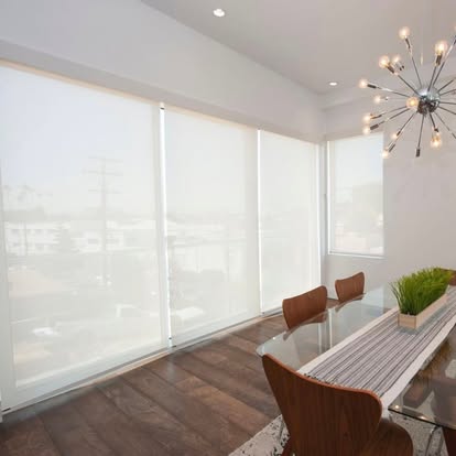 White solar roller shades across a wall of dining room windows with a sputnik chandelier and glass table