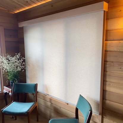 Light-filtering roller shade covering a wide window in a warm wood-paneled room with two chairs