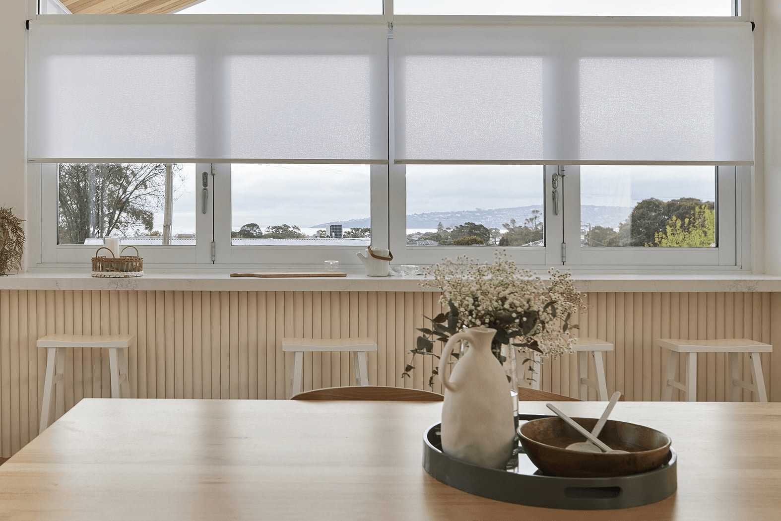 Four white light-filtering Soluna roller shades lowered over a bank of dining-room windows framing a coastal sea-and-hillside view