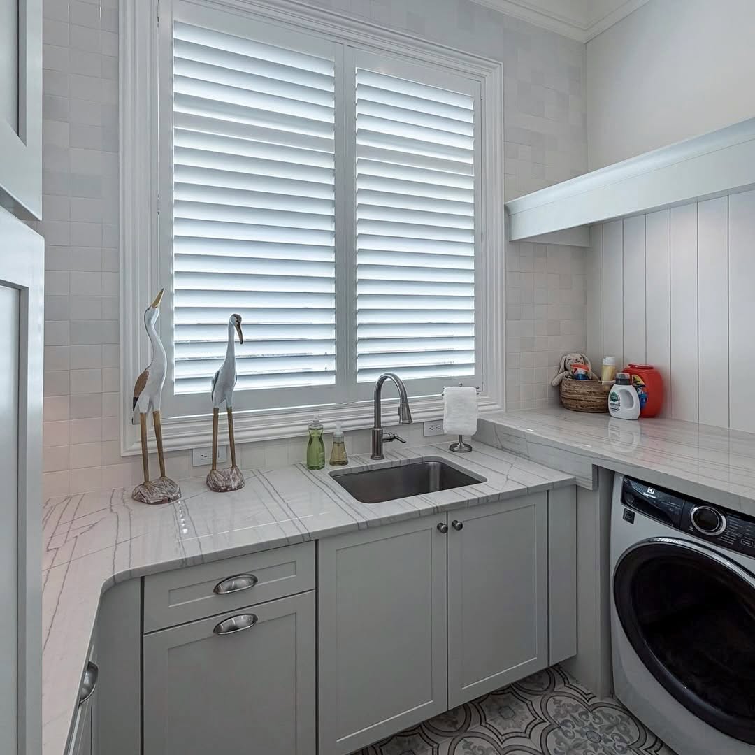 White Woodlore plantation shutters on a laundry-room window above a marble-topped sink with gray cabinets