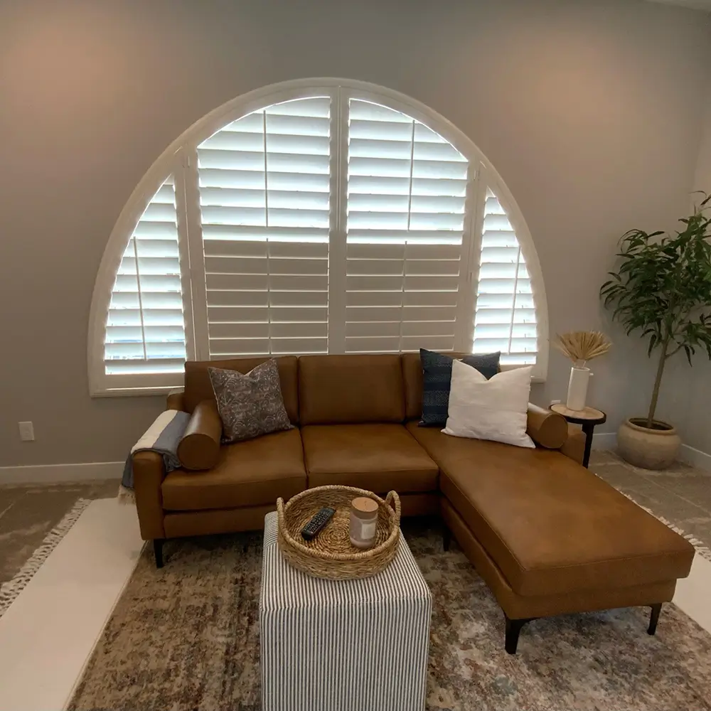 White Woodlore Plus plantation shutters fitted to a large arched window above a caramel leather sectional in a warm neutral living room
