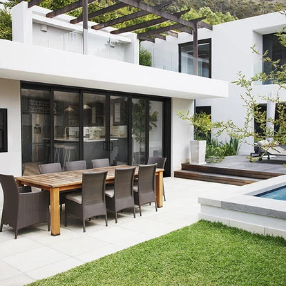 Modern white home with a pergola-shaded patio and black-framed PGT EnergyVue vinyl sliding glass doors opening to an outdoor dining area beside a pool