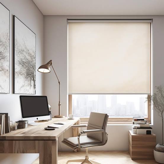 Cream Basix roller shade lowered over a large home-office window with a city view, above a wood desk and task chair
