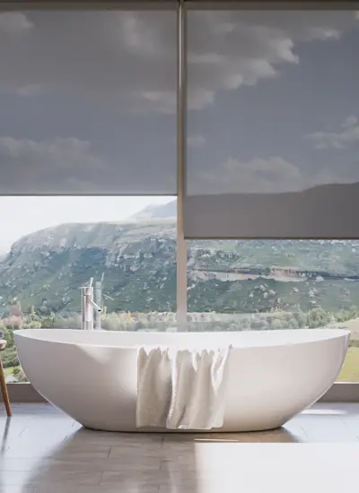 Soft gray COLOR roller shades lowered over floor-to-ceiling bathroom windows framing a green mountain valley, above a freestanding white soaking tub