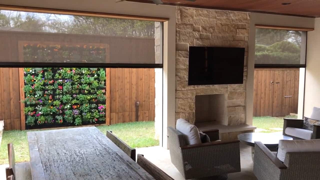 Dark Texton retractable exterior screens lowered over two openings of a covered patio with a stone fireplace and a colorful living plant wall beyond