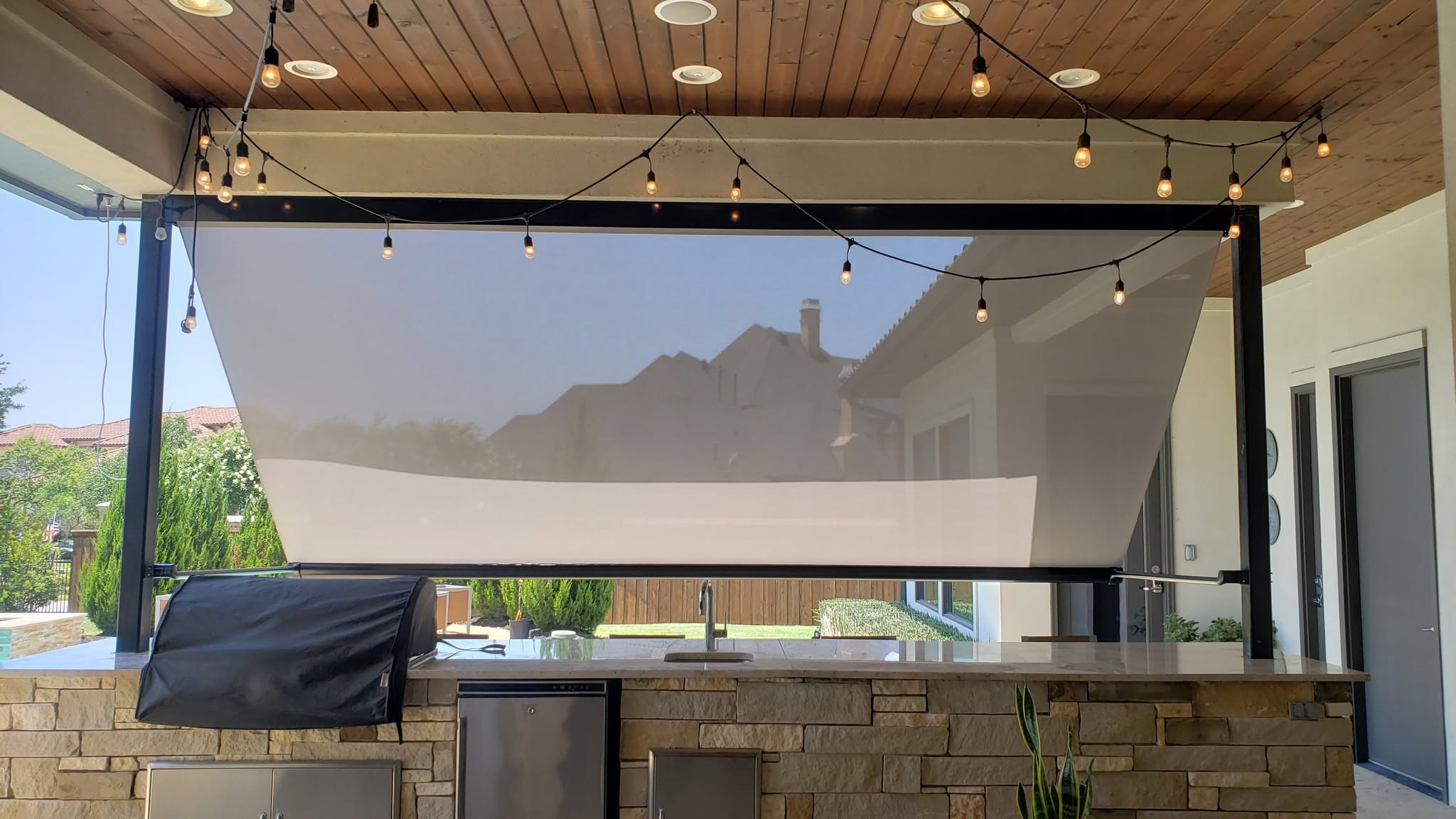 Texton pivot-arm retractable exterior screen angled outward over a stone outdoor kitchen with a built-in grill and string lights