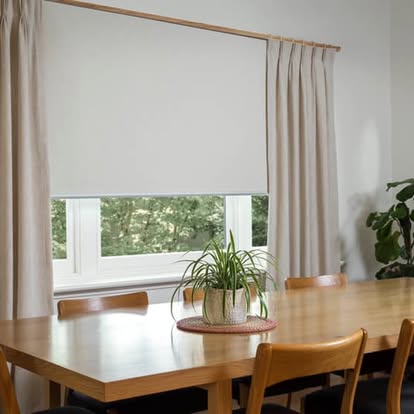White roller shade lowered over a window in a bright dining room with wood table and chairs